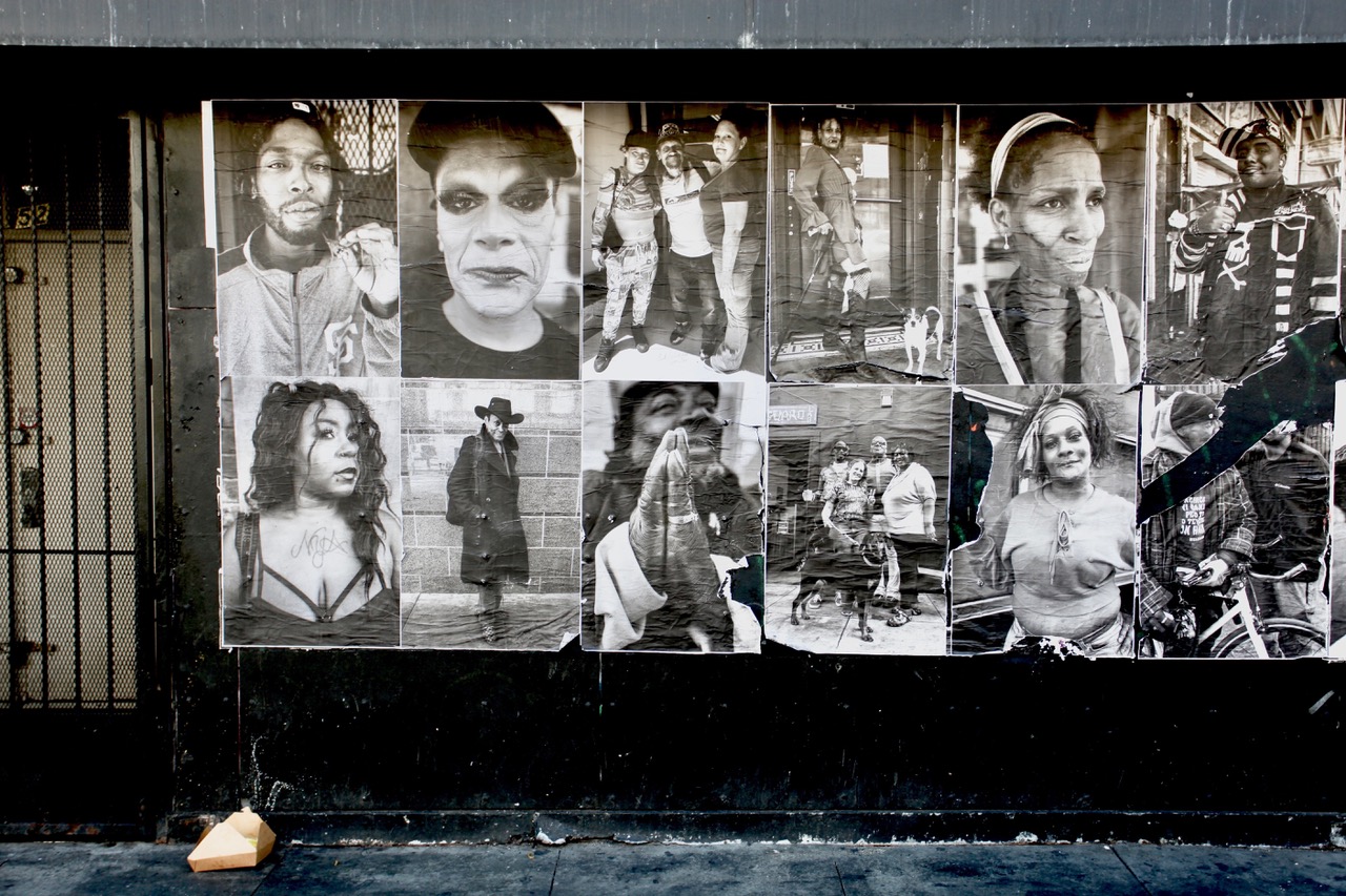 A wall displays a collage of black and white portraits of various people, each with distinct expressions and styles, in an urban setting.