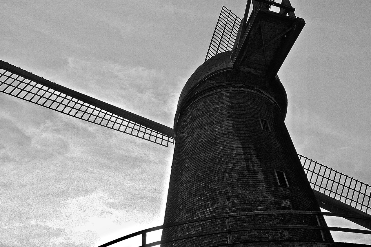 Black and white photo of a brick windmill viewed from below, showing its tower and large blades extending outward against the sky.