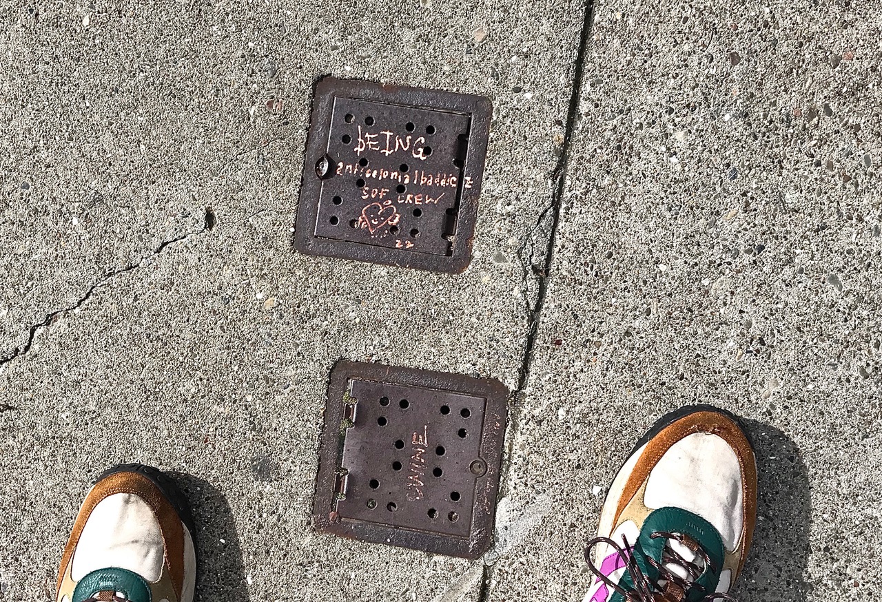 Two metal utility covers on a concrete sidewalk, one inscribed with graffiti, with parts of two shoes visible at the bottom corners of the image.