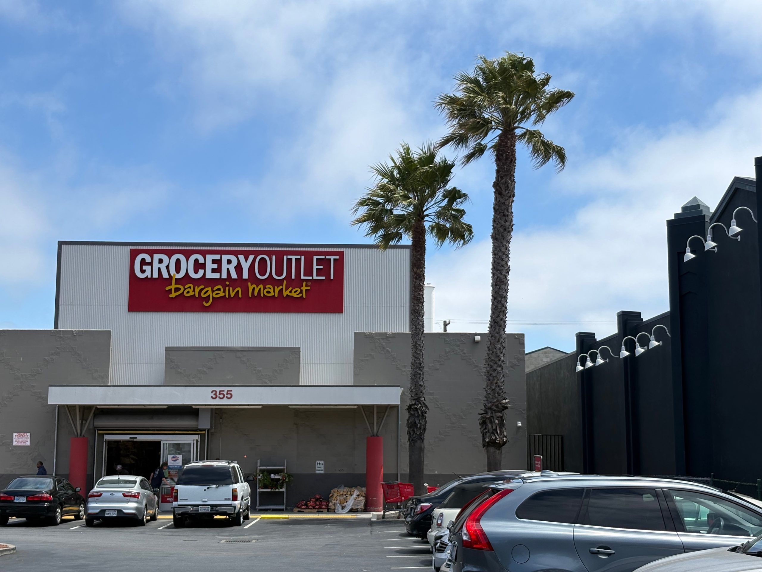 Grocery Outlet Bargain Market storefront with parked cars in front and two tall palm trees beside the entrance under a partly cloudy sky.