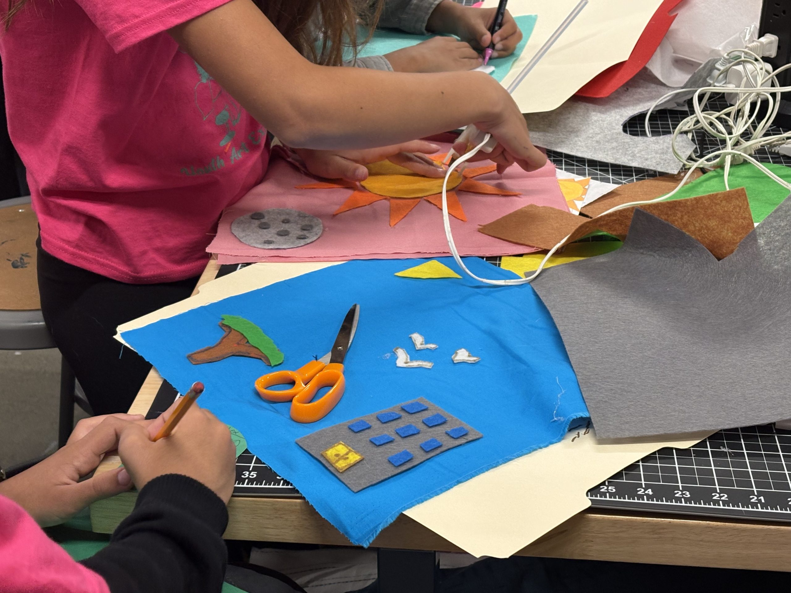 Children use scissors, pencils, and fabric pieces to create colorful felt art projects on a table covered with crafting tools and materials.