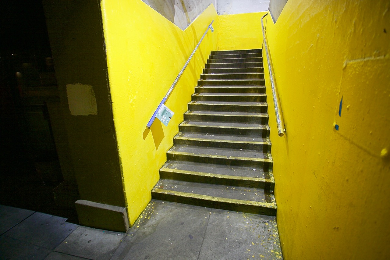 Concrete staircase with yellow-painted walls, metal handrails, and a few scattered pieces of litter at the base, leading upward to an unseen exit.