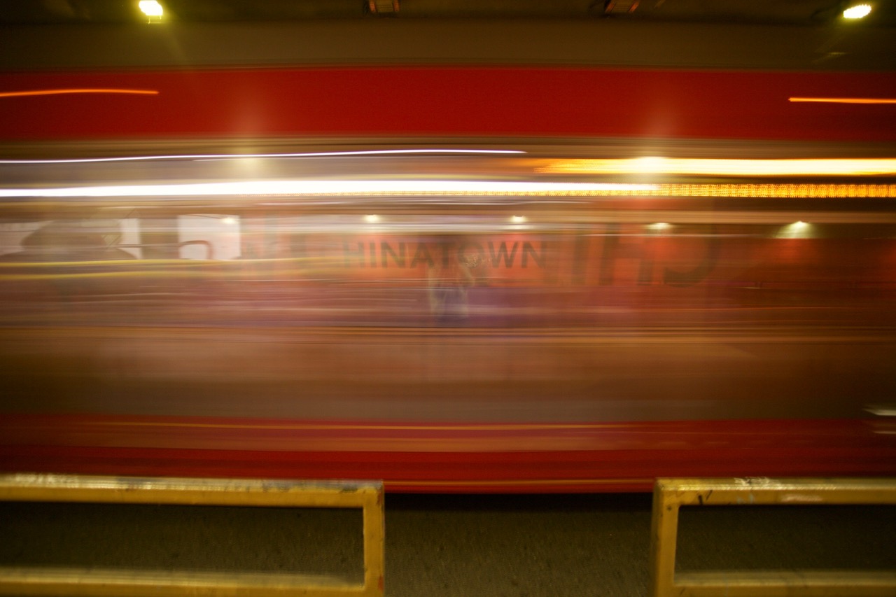 A red bus moves quickly past a street barrier, creating a motion blur effect; the word "Chinatown" is faintly visible through the blur.