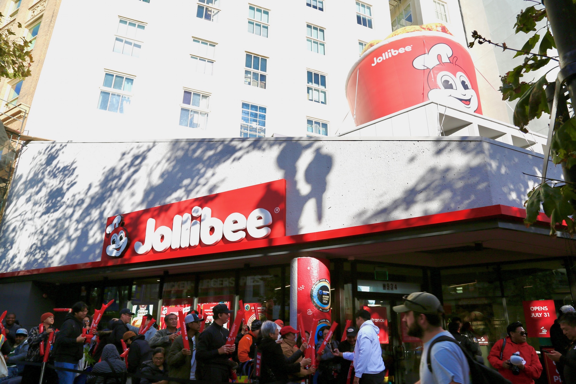 People stand outside a Jollibee restaurant with the logo and a giant bucket featuring the Jollibee mascot on the roof.