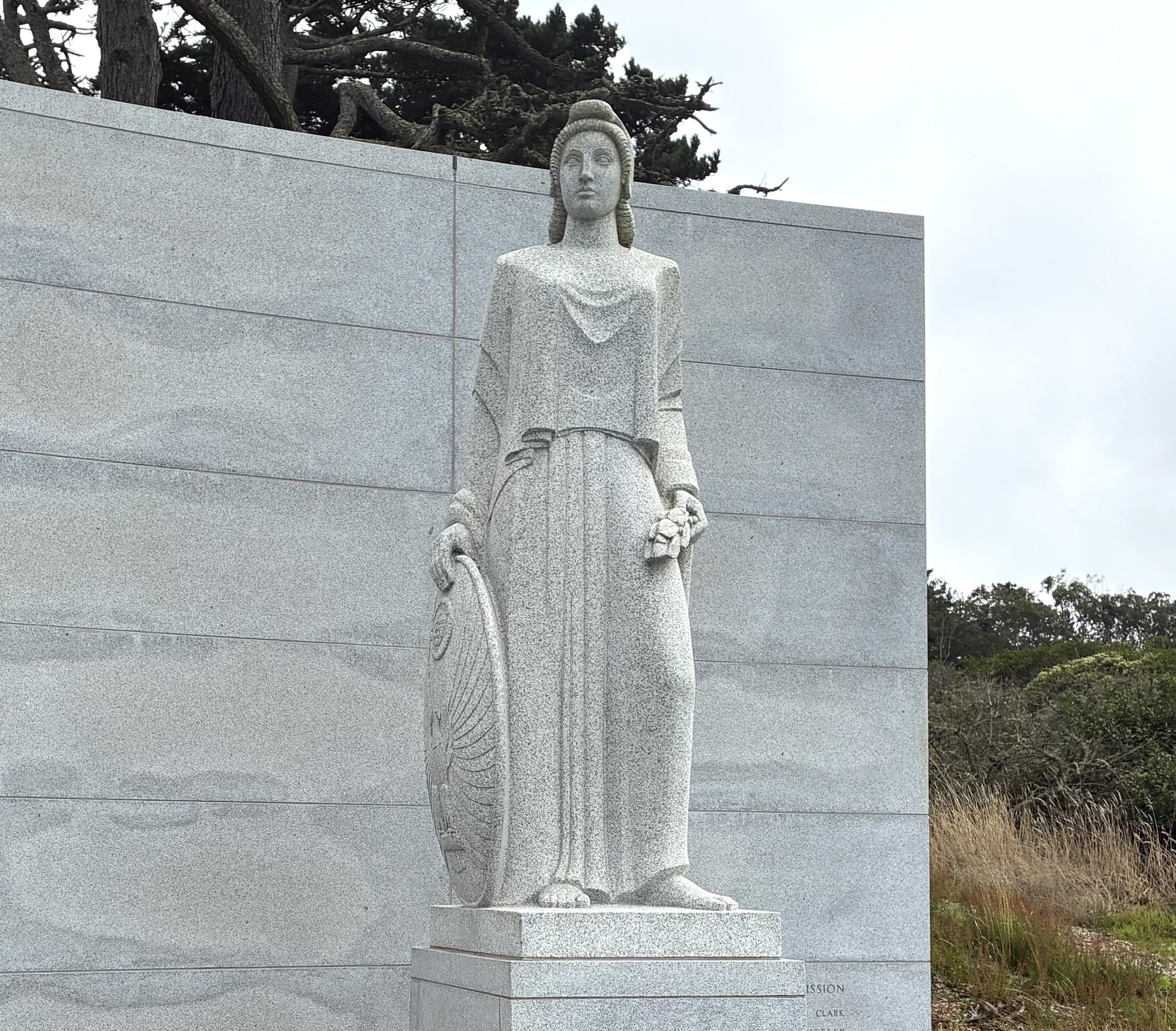 Granite statue of a woman holding a shield in her left hand and a laurel branch in her right, standing against a stone wall with trees in the background.