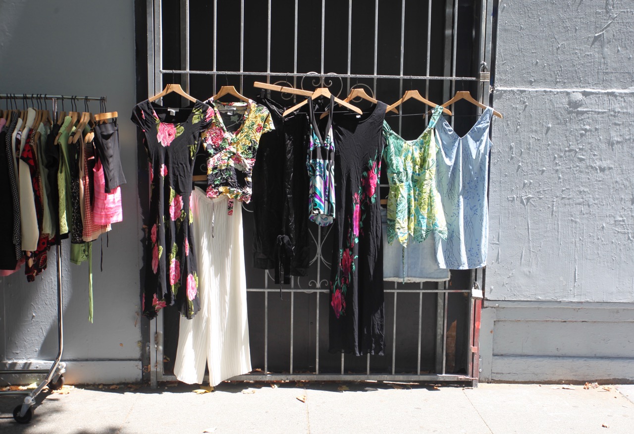 Various dresses and tops on hangers are displayed on a metal gate and clothing rack outdoors, with sunlight casting shadows on the sidewalk.