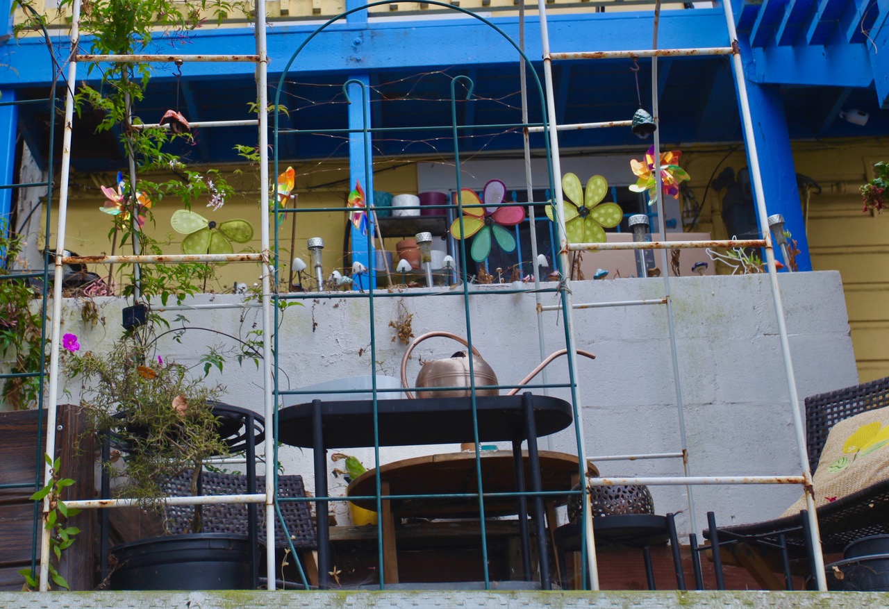 A patio with a table, chairs, watering can, potted plants, and colorful flower decorations against a concrete wall and blue railing.