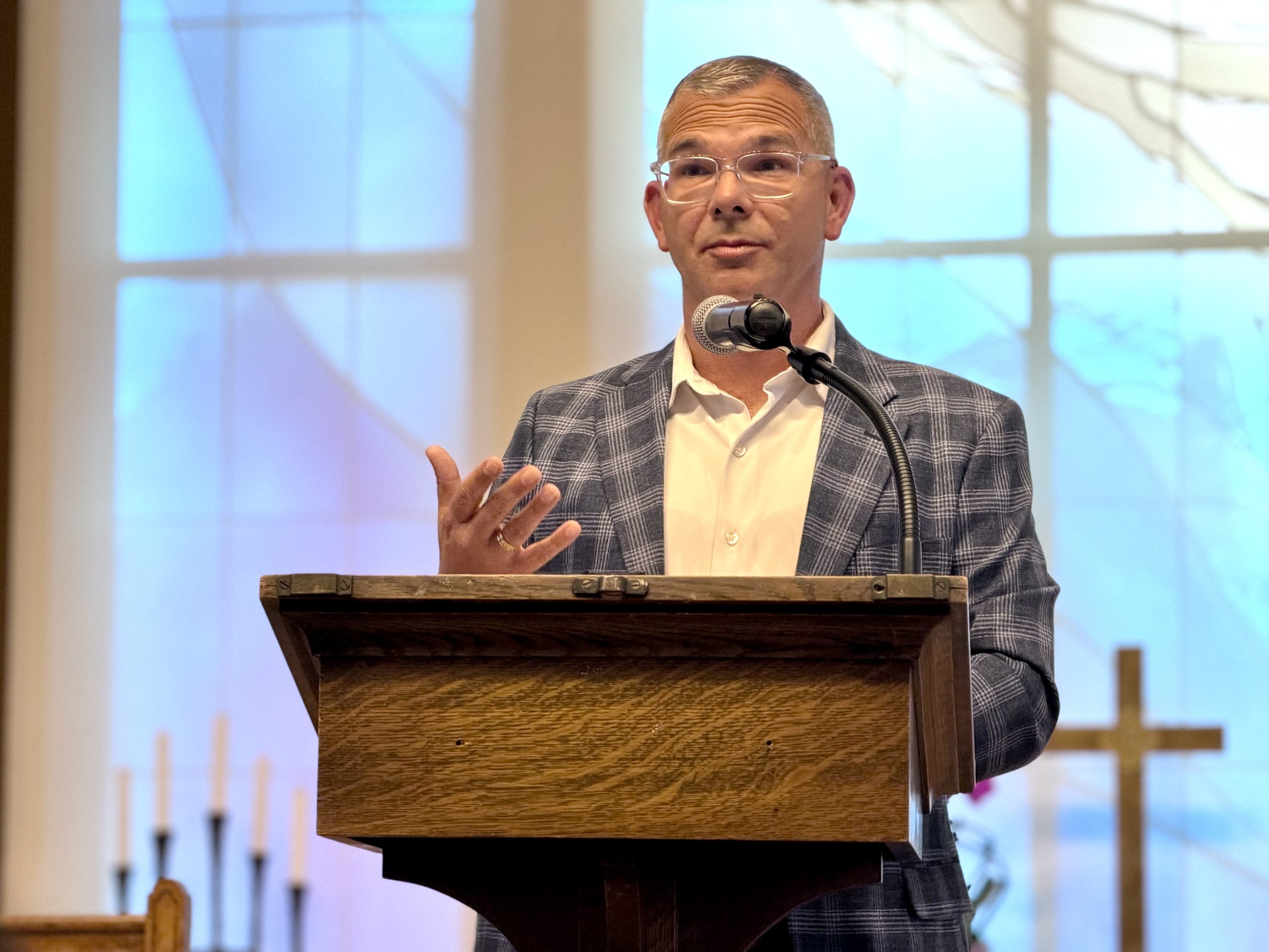 A man wearing glasses speaks at a podium in a church, with candles and a cross visible in the background.