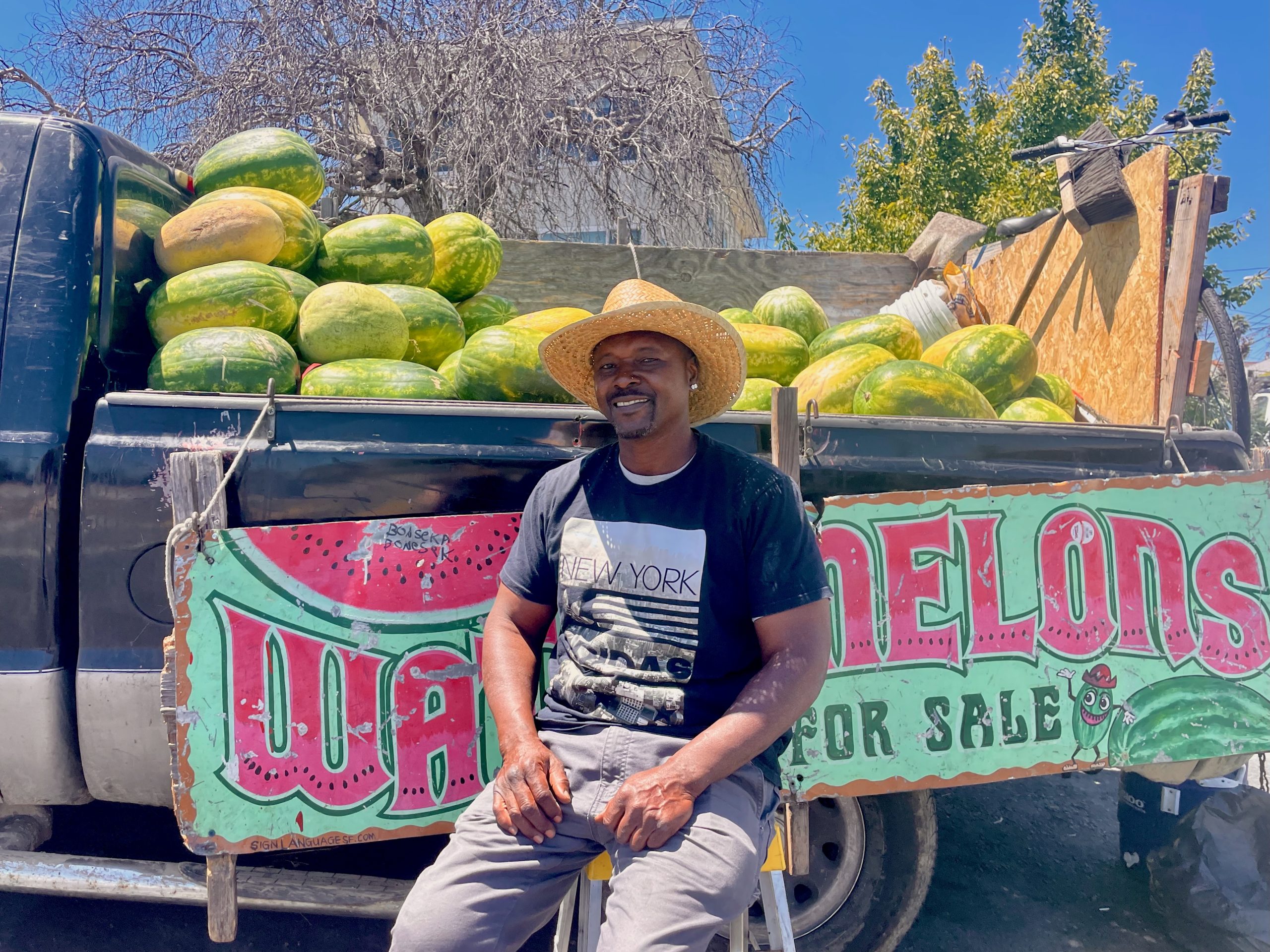 A man in a straw hat sits in front of a truck filled with watermelons and a “WATERMELONS FOR SALE” sign on a sunny day.