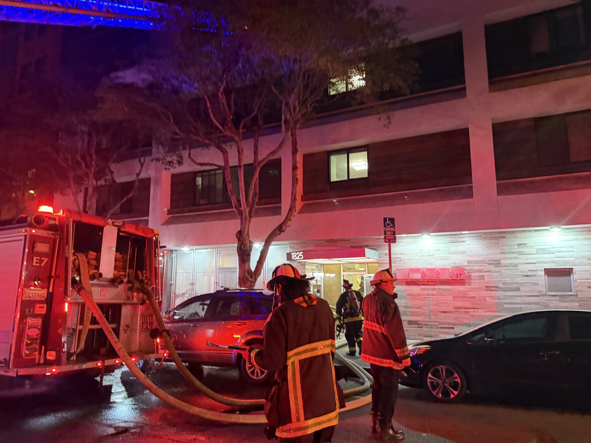 Firefighters stand outside a building at night with hoses from a fire truck extended into the entrance; emergency lights illuminate the scene.