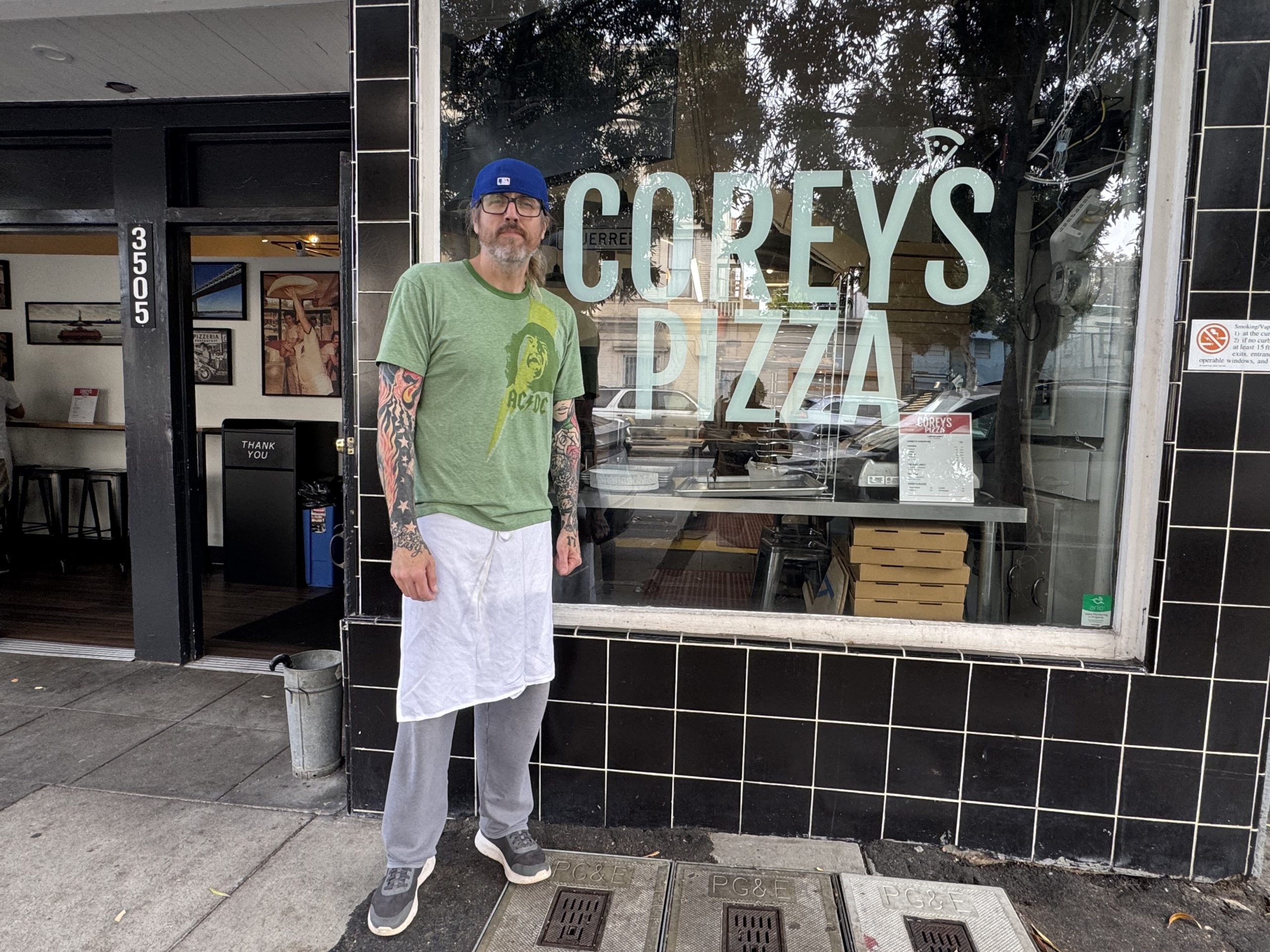 A man in a green T-shirt and white apron stands outside Corey’s Pizza, with stacked pizza boxes visible through the front window.