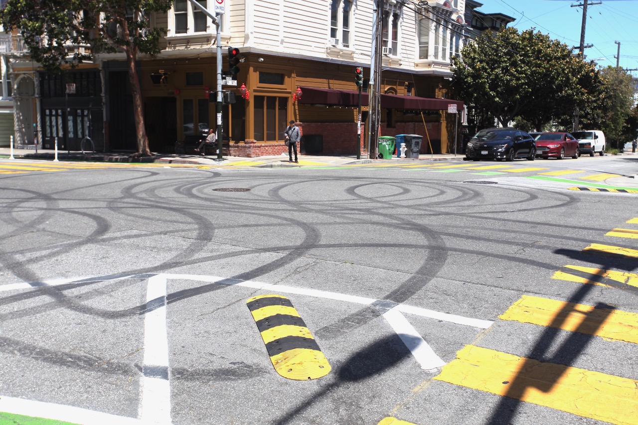 A city street intersection with tire skid marks on the pavement, traffic signals, parked cars, and a pedestrian walking on the sidewalk.