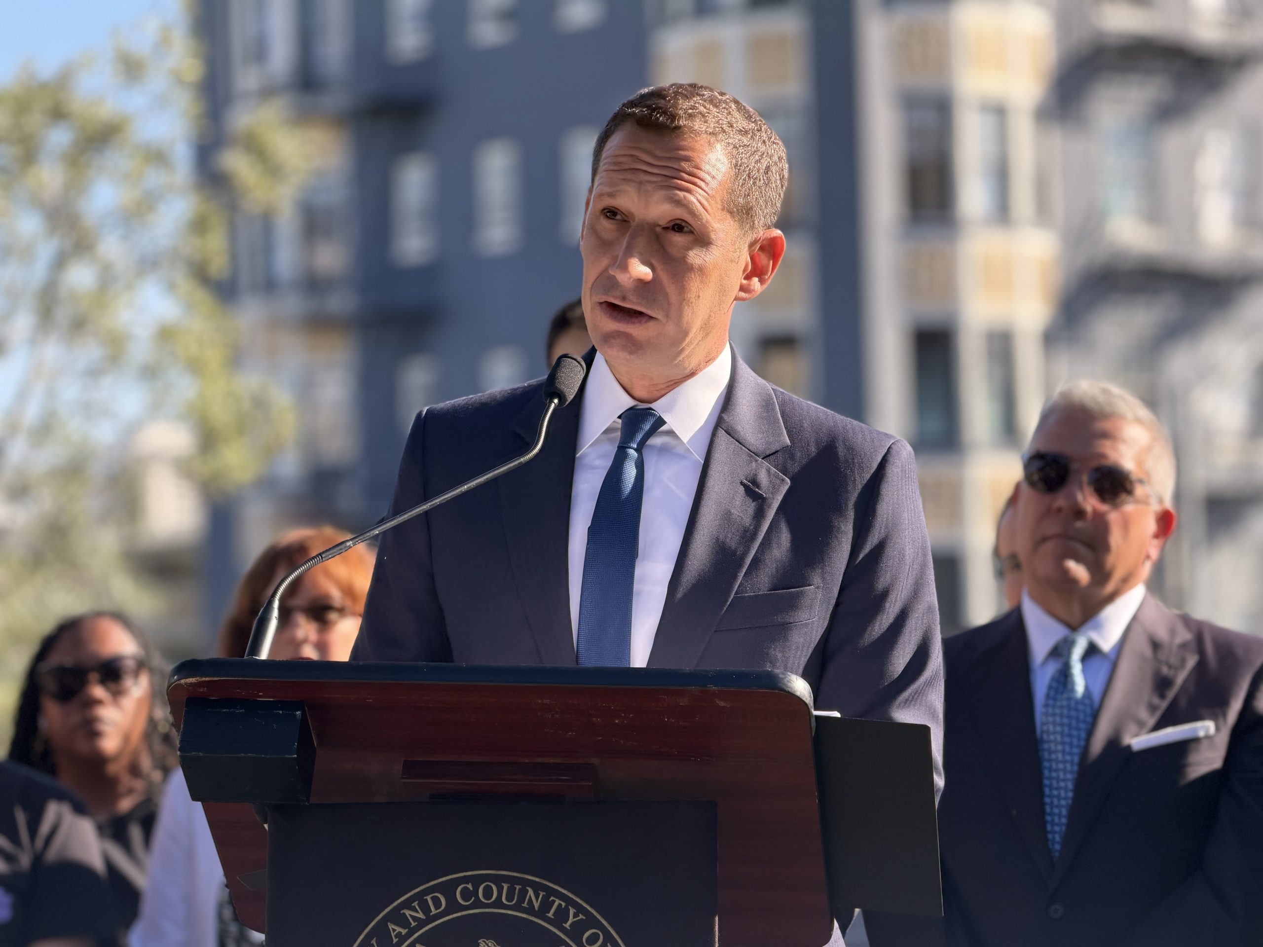 A man in a suit speaks at a podium outdoors, with several people standing behind him and apartment buildings in the background.