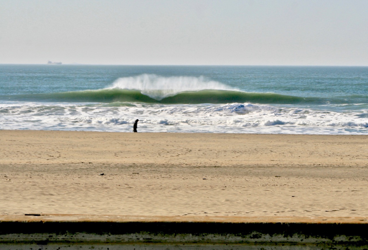 A person stands alone on a sandy beach with ocean waves breaking in the background and a ship visible on the horizon.
