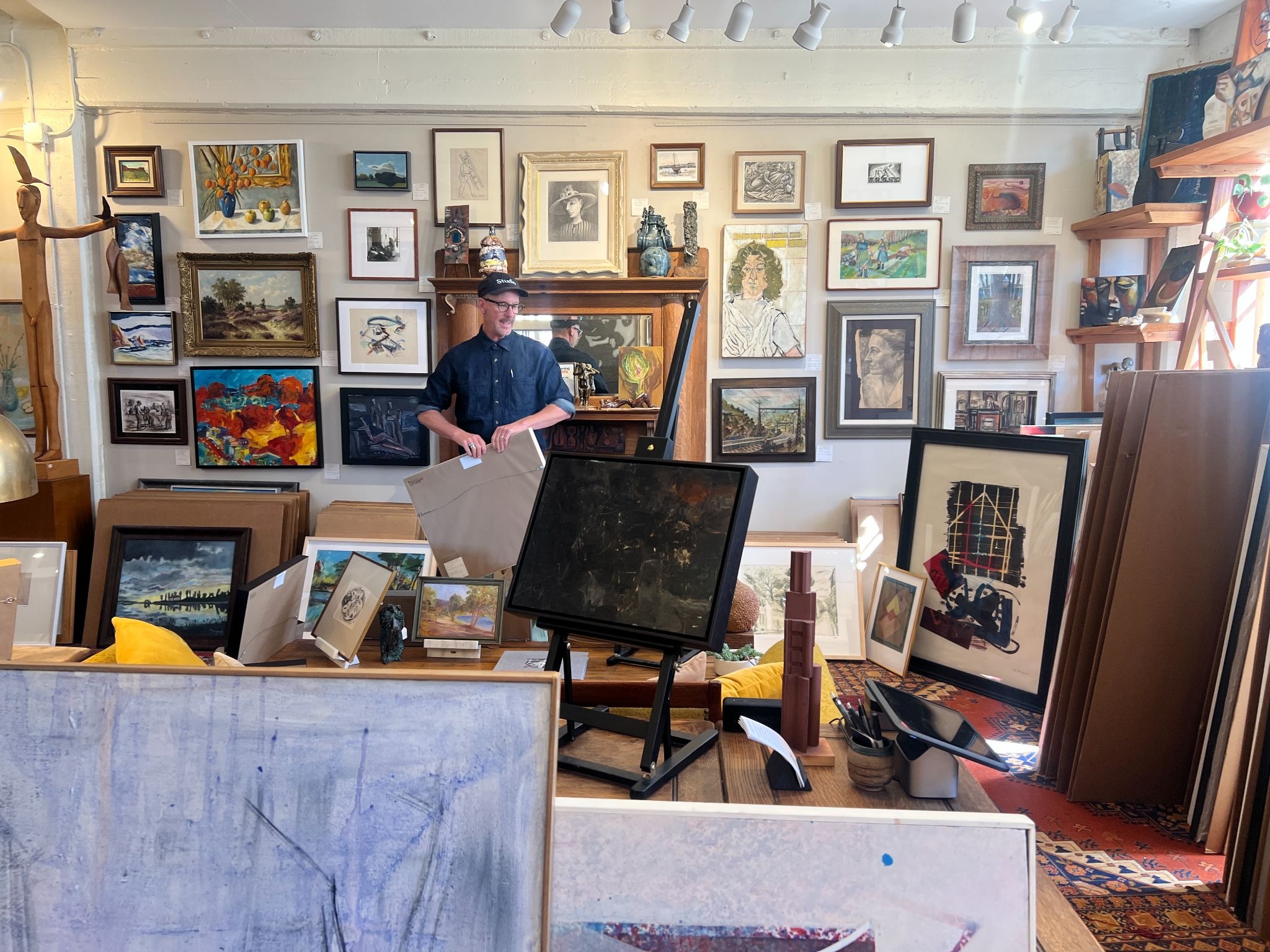 A man in a workshop holding a framed piece of art surrounded by numerous paintings on walls and easels.