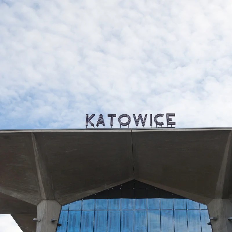 Indgangspartiet til Katowice togstation med moderne betonfacade og byens navn på taget mod blå himmel