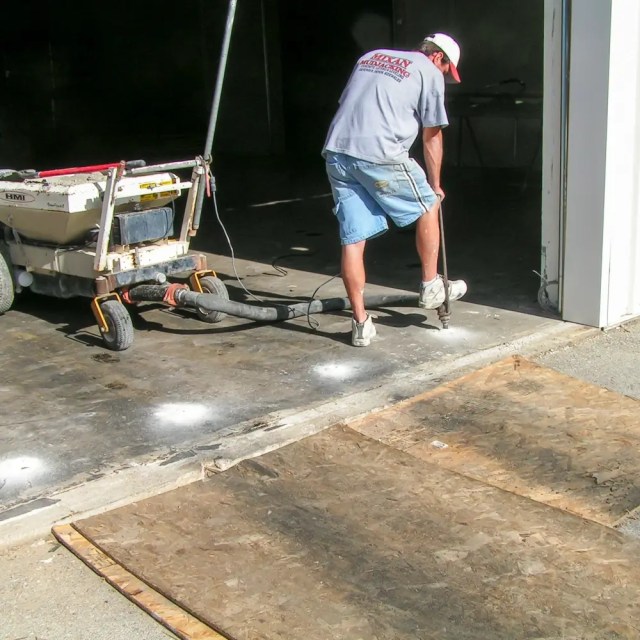 A worker in shorts and a T-shirt uses a long tool to fix or level a concrete garage floor near an open garage door, with machinery and plywood sheets visible nearby. - Mixan Mudjacking A worker in shorts and a T-shirt uses a long tool to fix or level a concrete garage floor near an open garage door, with machinery and plywood sheets visible nearby.