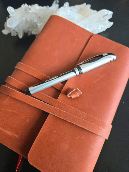 A silver pen rests on a closed, brown leather journal with a wraparound strap, suggesting a space for thoughtful reflection or perhaps jotting down tarot rules. Clear quartz crystals are visible in the background on a dark surface.