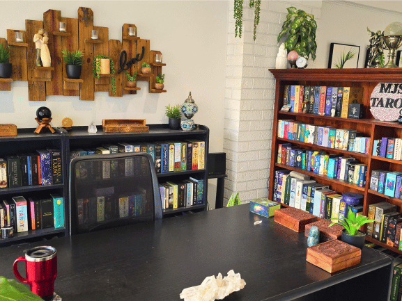 Home office with bookshelves full of  tarot decks, a black desk adorned with crystals and plants, a mesh chair, and a wall shelf holding small plants and figurines.