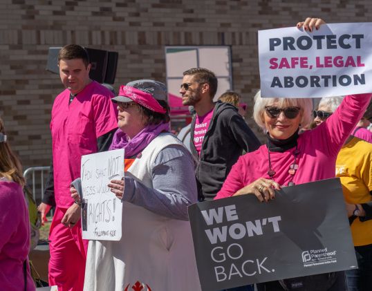 Photo of pro-choice protesters, one holding a sign that says "Protect Safe, Legal Abortion".