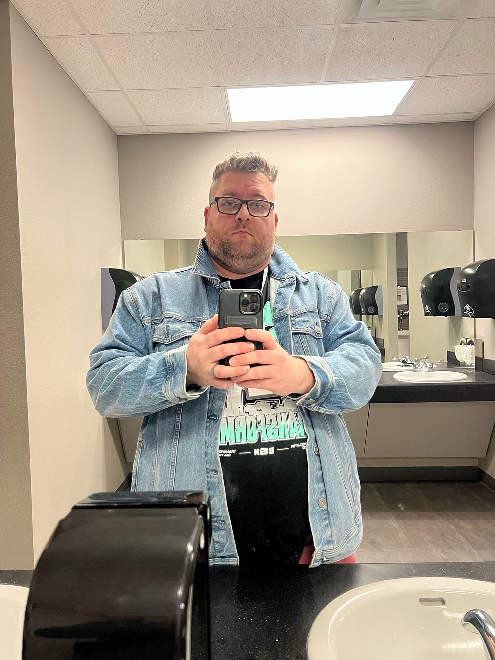 A person taking a selfie in a restroom mirror, wearing glasses and a denim jacket over a black t-shirt with graphic design.