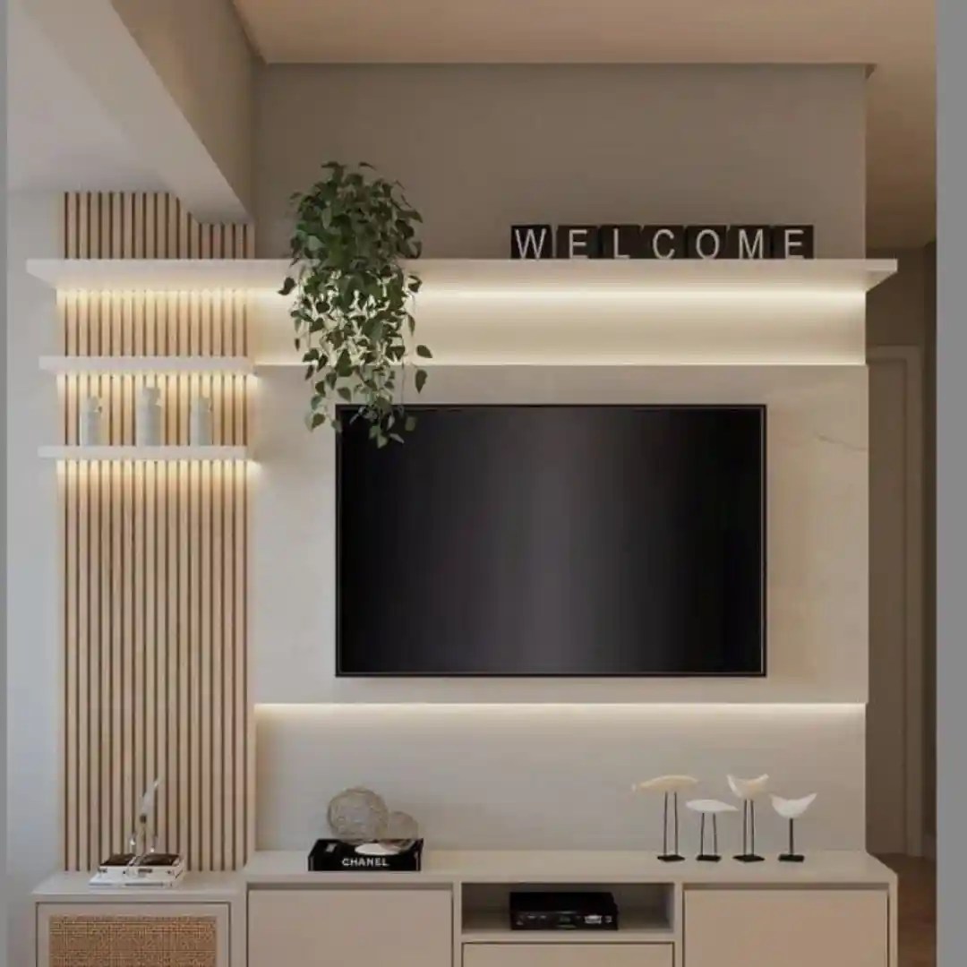Modern living room TV wall with wooden slats, LED lighting, floating shelves, and "WELCOME" sign. Minimalist design with green plant and decor.