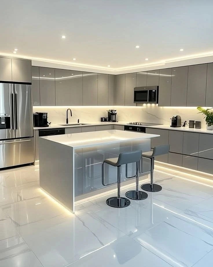 Sleek modern kitchen with gray cabinets, white marble floors, a large island, and under-cabinet lighting. Stainless steel appliances and bar stools complete the look.