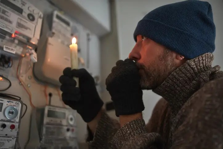 Man bundled in winter clothing holding a candle near an electric meter, symbolizing rising energy costs and the need to protect your finances from inflation.