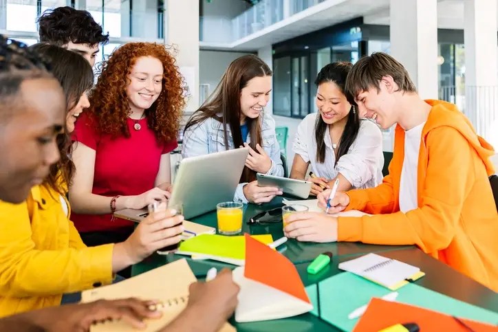 Group of students collaborating on a project about financial education in schools using laptops, tablets, and notebooks at cafeteria campus table.
