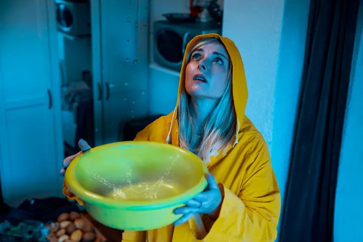 Woman in a raincoat catching water from a ceiling leak, symbolizing the need for emergency preparedness financial planning during unexpected disasters.