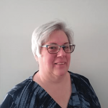 Smiling woman with short gray hair and glasses, wearing a blue patterned blouse, standing against a light gray wall.