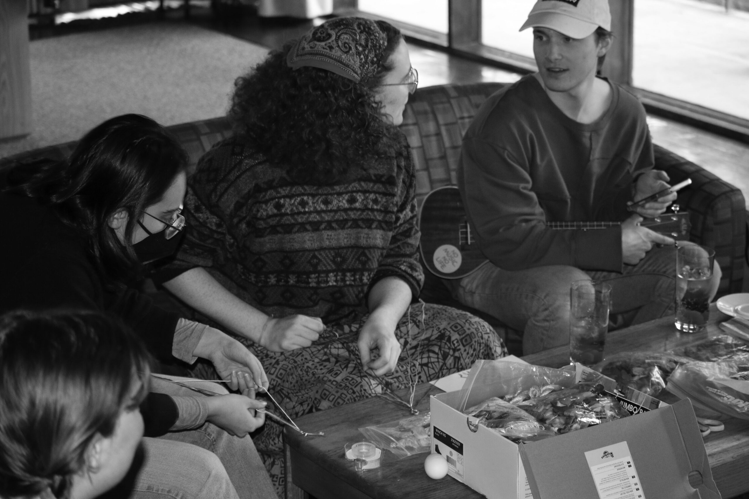 Photo of two club members talking, one with a ukulele, while another works on a friendship bracelet