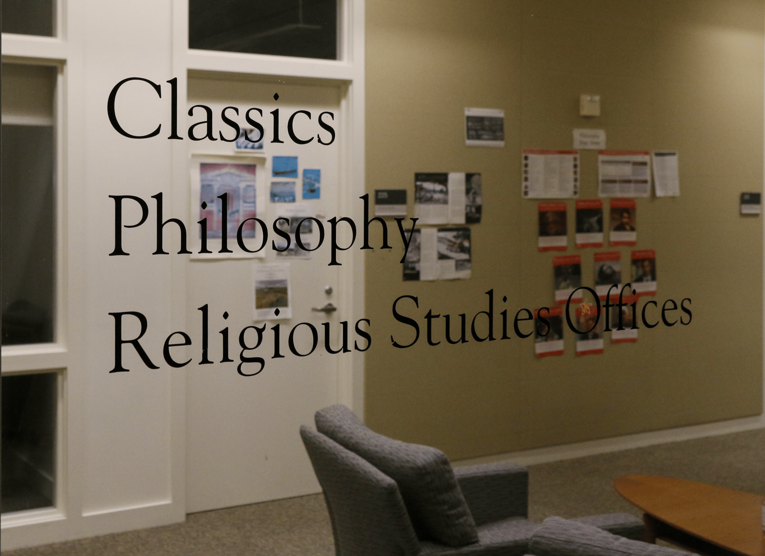 Sign for Classics, Philosophy and Religious Studies offices labeled on glass. Through the glass, while blurry, an office door surrounded by small windows and a wall with diagrams can be seen.