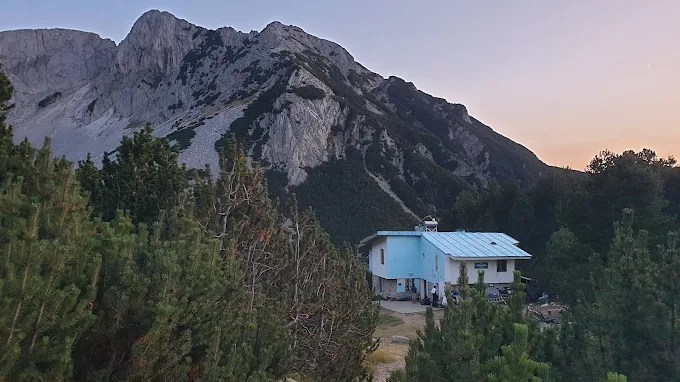 Blue-roofed mountain hut below a grey rock peak at dusk