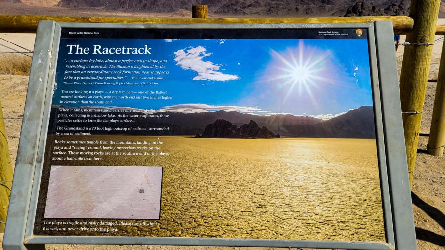 A photo of a sign post that says The Racetrack in white letters on top of a photo of a desert landscape with a white sun. Visiting Racetrack Playa in Death Valley. MPA Project Travels.
