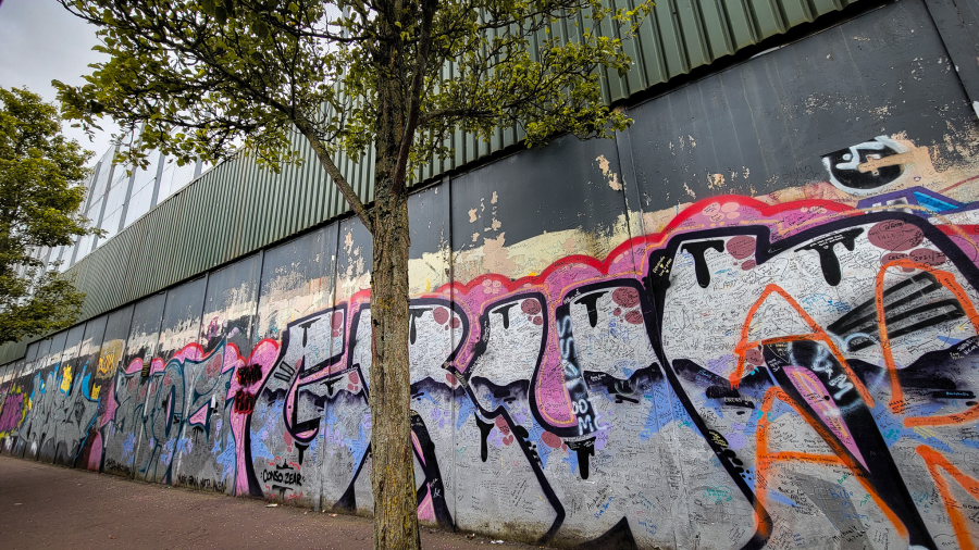 A white, pink, and black mural in graffiti on a large wall. Lots of signatures on the white paint of the mural. Black Cab Tour. 24 Hours in Belfast. MPA Project Travels.