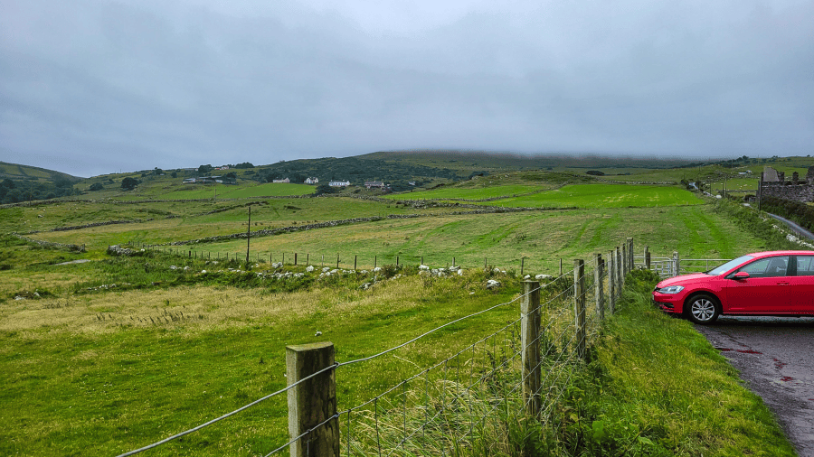 A red car sits on the righthand side of a photo of green fields and a cloudy sky. Renting a car in Ireland. MPA Project Travels.