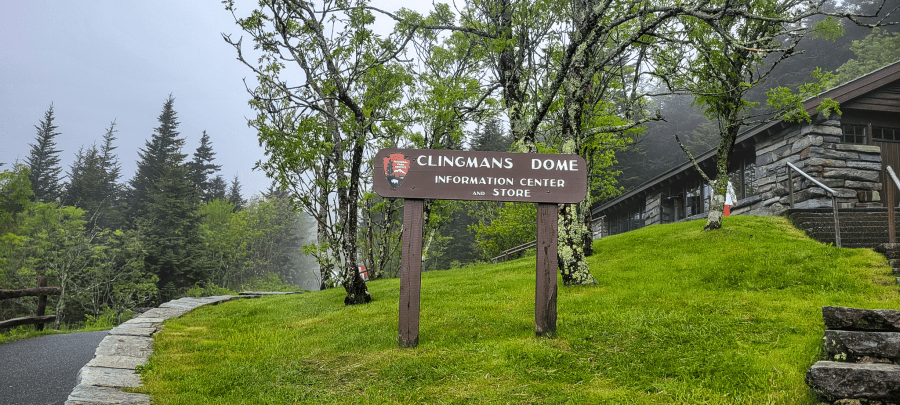 Kuwohi (Clingmans Dome) information center sign, surrounded by green grass and trees in fog.