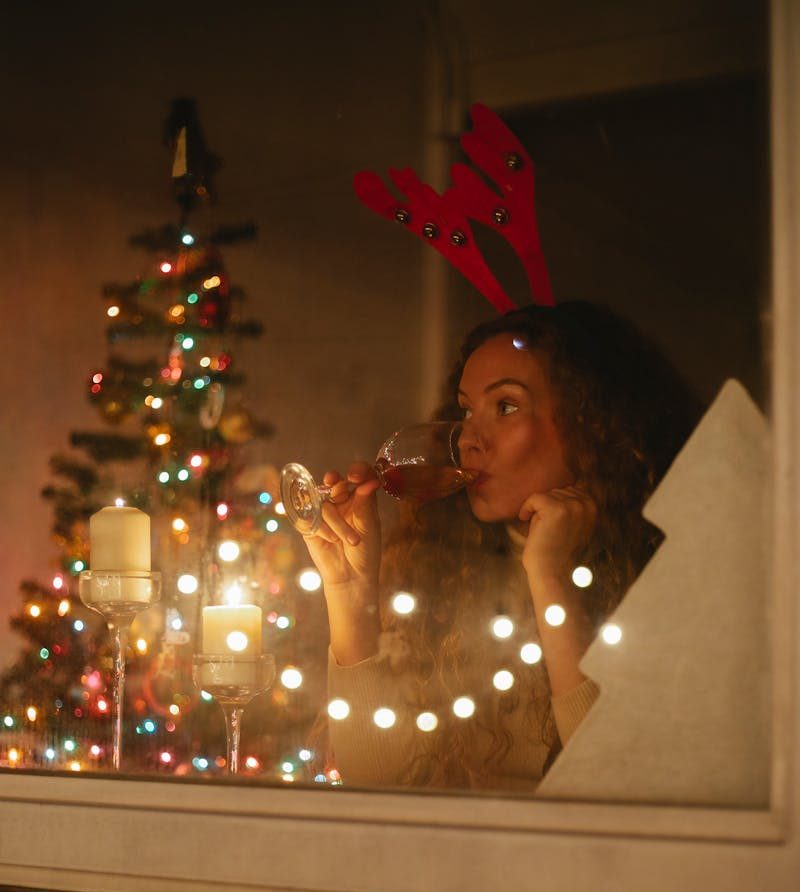 Through glass side view of contemplative female enjoying alcoholic beverage against decorated fir tree with shiny garland while looking away