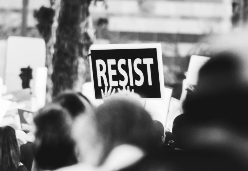 Monochrome image showing a crowd holding a 'Resist' sign at a protest.