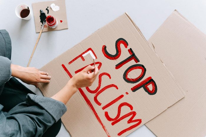 Person creating protest sign with "Stop Fascism" message, using red and black paint.