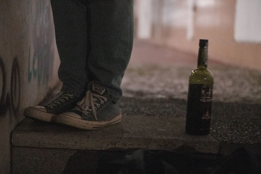 Legs in sneakers next to a wine bottle on urban steps in low light setting.