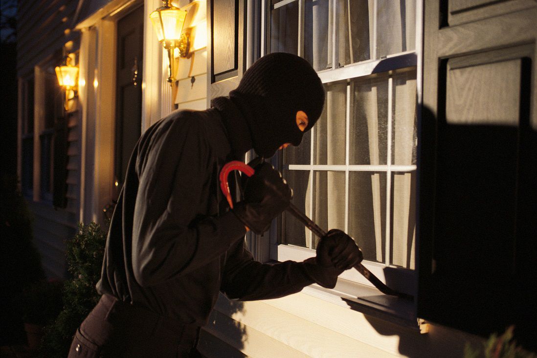 A person wearing a balaclava and dark clothing is using a crowbar to pry open a window of a house at dusk, with outdoor lights on either side of the entrance.
