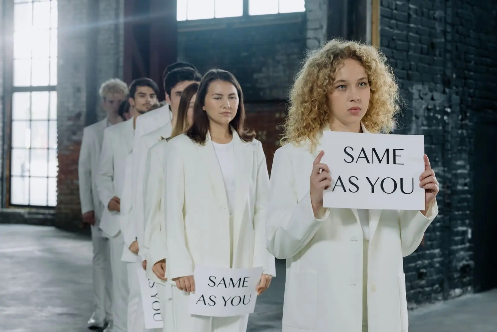 People dressed in white standing in a line, each holding an identical sign that reads "Same as you"