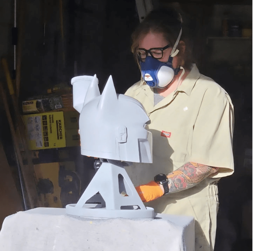 Kristie working on a batman helmet, an example for the 3d printing portfolio. The mask is unpainted and on a stand on a material covered work bench. Krisite is wearing a protective mask and is wearing workclothes.