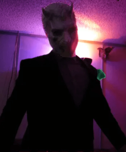 Person in a devil mask and black suit posing in a dimly lit room with purple and pink lighting.