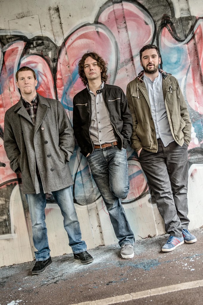 Three men posing against a colorful graffiti backdrop, wearing casual clothing and standing in a relaxed posture.
