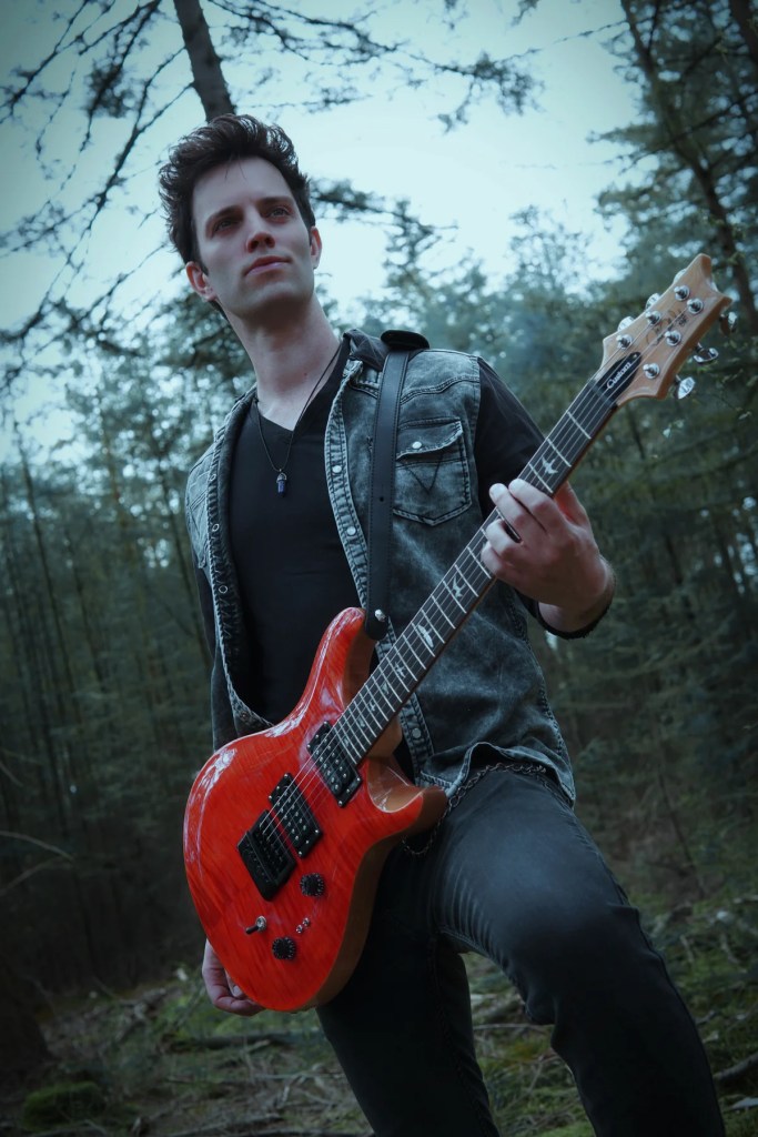A young man playing a red electric guitar in a forest setting, wearing a denim vest and black shirt.