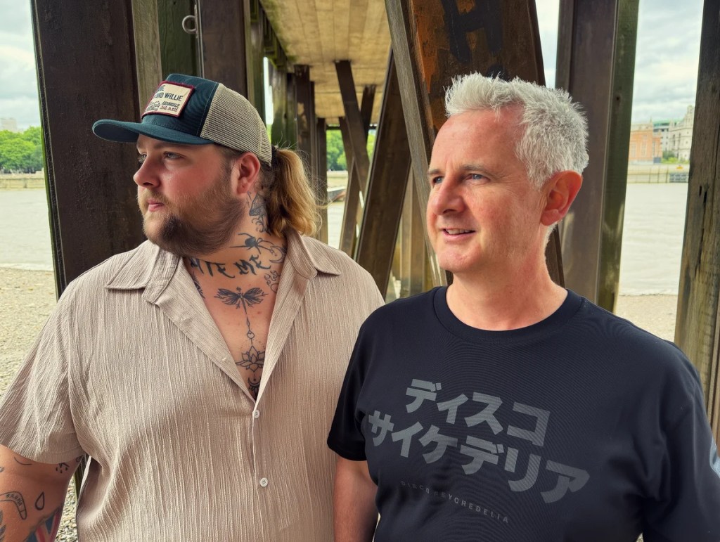 Two men standing under a wooden structure by a river. One man, with a ponytail and tattoos, wears a light-colored button-up shirt and a cap. The other man, with short gray hair, wears a dark t-shirt and is smiling.