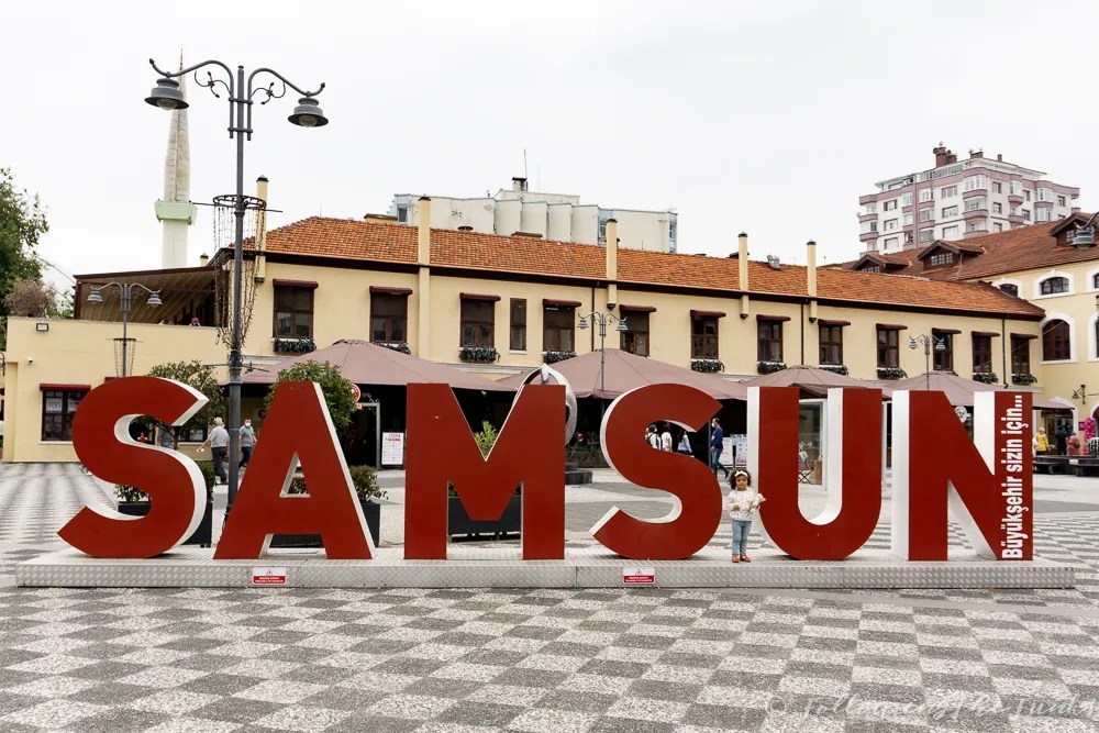 Samsun yazılı büyük harflerin önünde bir çocuk, şehir meydanında yer alan tarihi binalar ve çiçeklerle çevrili.